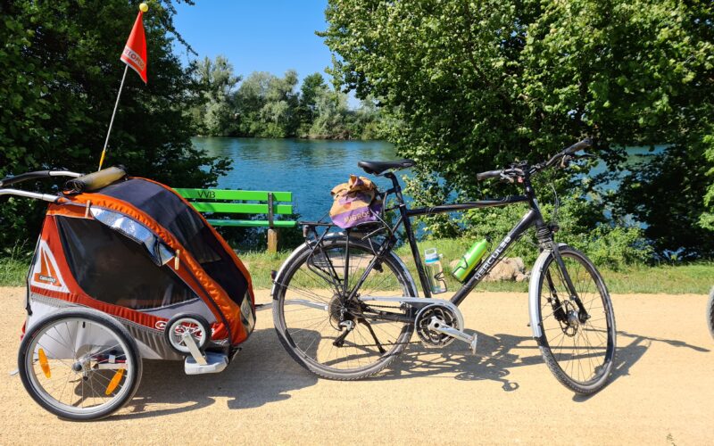 Rimorchietto da bici con bambini e bicicletta davanti alla riva dell’Aare, con un braciere e una panchina