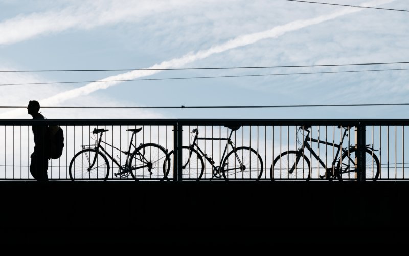 Silhouette eines Mannes mit einem Rucksack, der an drei Fahrrädern vorbeigeht, die an ein Brückengeländer gekettet sind.